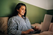 Freelancer feminino de meia-idade positivo com cabelos longos e escuros em roupas casuais sentadas na cama e trabalhando remotamente no laptop em casa — Fotografia de Stock
