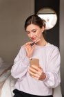Щаслива молода жінка-блогер в повсякденному одязі посміхається і демонструє блиск губ під час зйомок відео на смартфоні для відеоблогу — стокове фото
