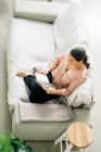 Dall'alto corpo pieno di libro di lettura femminile scalzo mentre seduto su comodo divano in soggiorno con pianta verde a casa — Foto stock