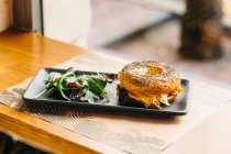 Sanduíche de bagel apetitoso com queijo e frango servido em prato com salada de rúcula na mesa no café — Fotografia de Stock