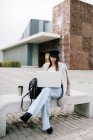 Jeune entrepreneure concentrée en tenue tendance assise sur un banc et parcourant un ordinateur portable tout en travaillant sur un projet à distance en ville — Photo de stock