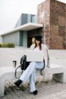 Concentrated young female entrepreneur in trendy outfit sitting on bench and browsing laptop while working on remote project in city — Stock Photo