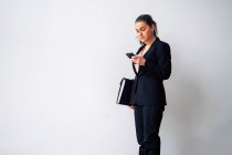 Серьёзная предпринимательница средних лет с хвостиком в чёрном костюме и текстовыми сообщениями на мобильном телефоне, стоя на белом фоне с папкой — стоковое фото