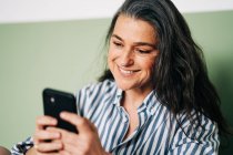 Позитивна жінка середнього віку з довгим темним волоссям в повсякденному одязі посміхається під час розмови на мобільному телефоні, сидячи на ліжку вдома. — стокове фото