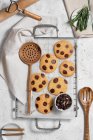 Draufsicht auf frisch gebackene süße Kekse mit Schokoladenstücken auf Metallgitter auf dem Tisch mit verschiedenen Küchengeräten und grünen Rosmarinzweigen — Stockfoto