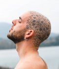 Боковой вид спокойного мужчины с татуированной лысой головой, стоящего с закрытыми глазами на природе против реки и горы в пасмурный день — стоковое фото