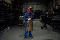 Anonymous mechanic in protective helmet and apron on blue overall standing with welding hose in light workshop near metal constructions — Stock Photo
