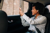 Side view of cheerful African American female smiling and talking in video chat in mobile phone in modern automobile — Stock Photo