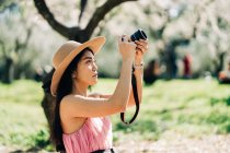 Seitenansicht einer fokussierten ethnischen Frau mit Strohhut, die ein Foto mit der Kamera im Garten mit blühenden Bäumen macht — Stockfoto