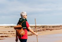 Side view of elderly female backpacker with trekking pole strolling on boulders against stormy ocean under cloudy sky — Stock Photo