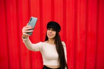 Enthousiaste femme entrepreneure branchée debout contre le mur rouge en ville et prendre soi-même tir sur smartphone — Photo de stock