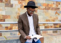 African American male in stylish hat and jacket standing on city street and reading interesting book — Stock Photo
