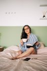 Donna intelligente negli occhiali con i capelli lunghi e notebook che distoglie lo sguardo e poggia su un morbido letto in camera da letto — Foto stock