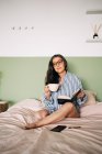 Kluge Frau mit Brille, langen Haaren und Notizbuch schaut weg und ruht sich auf weichem Bett im Schlafzimmer aus — Stockfoto