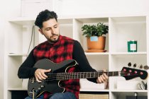 Vollbärtiger erwachsener Musiker spielt Bassgitarre während der Probe gegen Regale zu Hause — Stockfoto