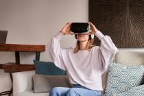 Вміст молодої жінки в повсякденному вбранні дивитися відео за допомогою VR окулярів, сидячи на зручному дивані вдома — стокове фото