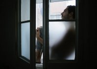 Vista lateral de las rodillas masculinas pensativas abrazando y sentado en el alféizar de la ventana en casa - foto de stock