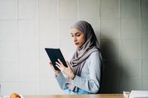 Vista lateral de la mujer musulmana freelancer usando hijab tradicional de pie en la cafetería y la tableta de navegación mientras trabaja en el proyecto de forma remota - foto de stock
