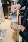 Desde arriba de cultivo barista anónimo verter leche en la taza de café mientras se hace arte latte en la cafetería - foto de stock
