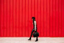 Вид збоку на жінку-підприємця в модному одязі, що йде вулицею на фоні червоної стіни і дивиться в сторону — стокове фото