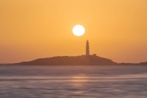 Cenário pitoresco de ondulação de água da costa de lavagem do oceano com farol de elevação colocado em Faro de Trafalgar em Cádiz, na Espanha, sob o céu laranja brilhante ao nascer do sol — Fotografia de Stock