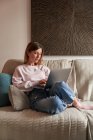 Full body of peaceful young female freelancer in casual clothes sitting on cozy sofa and working remotely on laptop — Stock Photo