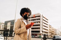 From below of stylish African American female in cloth mask texting message on mobile phone while walking on street of city — Stock Photo