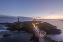 Spectacular scenery of bridge leading to rocky island covered with green grass with lighthouse placed in wavy ocean in Faro Illa Pancha in Galicia in Spain in twilight — Stock Photo