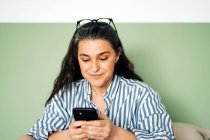 Positive middle aged Hispanic female with long dark hair in casual clothes smiling while messaging on mobile phone sitting on bed at home — Stock Photo