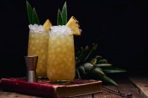 Wooden table with glasses of fresh yellow cocktails with ice cubes and pineapple pieces and leaves near spoon and shot glass placed on red book on black background — Stock Photo