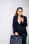 Emprendedora femenina seria de mediana edad con cola de caballo que usa mensajes de texto de traje negro en el teléfono celular mientras está de pie sobre fondo blanco con carpeta - foto de stock