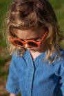 High angle of cute happy little girl in trendy clothes and sunglasses standing and relaxing on grassy lawn — Stock Photo