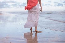 Вид ззаду анонімної жінки, що йде у хвилястій воді величезного океану на піщаному пляжі під хмарним небом — стокове фото