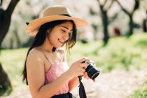 Side view of focused ethnic female in straw hat taking photo on camera in garden with blooming trees — Stock Photo