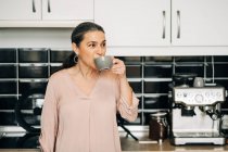 Allegro femmina di mezza età con tazza di bevanda calda in piedi al bancone della cucina con armadi bianchi e macchina da caffè moderna a casa — Foto stock