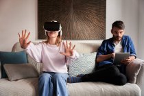 Positive junge Frau erlebt Virtual Reality in modernem Headset sitzt auf bequemem Sofa in der Nähe konzentrierter ethnischer Freund surft Tablet — Stockfoto