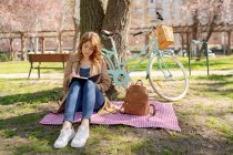 Corpo inteiro de jovem fêmea concentrada tomando notas no caderno em tecido quadriculado com mochila perto de bicicleta no parque — Fotografia de Stock