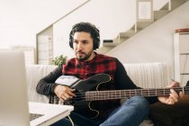 Adult male musician in headphones playing bass guitar against netbook on sofa in living room — Stock Photo