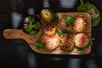 Vue du dessus de tomates noires fraîches mûres tranchées et de tiges de menthe verte sur planche à découper en bois sur fond noir — Photo de stock
