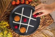 Top view of crop unrecognizable cook putting whole raspberry on plate with fritters and whipped cream representing noughts and crosses game — Stock Photo
