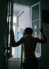 Visão traseira do macho com torso nu em pé perto da janela em casa e olhando para longe — Fotografia de Stock