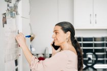 Side view of positive middle aged female making notes in calendar on fridge while having phone call in modern kitchen — Stock Photo