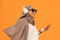 Вид сбоку искренней этнической женщины в платке и беспроводных наушниках с сотовым телефоном, слушающей музыку при дневном свете — стоковое фото