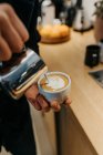 Зверху анонімний бариста наливає молоко в чашку кави, роблячи латте мистецтво в кафетерії — стокове фото
