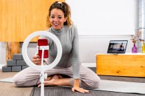 Ganzkörper positive erwachsene athletische Frau in Aktivkleidung, die ihr Smartphone am Selfie-Ring einrichtet, während sie auf dem Boden sitzt, bevor sie Vlog während des Yoga-Trainings filmt — Stockfoto