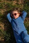 De cima menina em roupas da moda e óculos de sol de mãos dadas atrás da cabeça e relaxante no gramado gramado — Fotografia de Stock