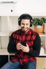 Adult bearded male musician in headphones text messaging on cellphone in living room — Stock Photo