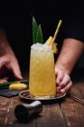 Анонимный бармен, собирающий спиртный коктейль с зелеными листьями и ананасом на подносе возле рюмки за деревянным столом на черном фоне — стоковое фото
