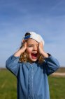 Menina loira feliz no chapéu tocando a cabeça e olhando para longe com a boca aberta contra o céu azul no verão no prado — Fotografia de Stock