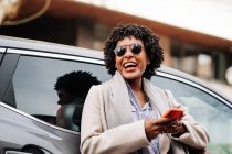 Низький кут сміху афро-американської жінки з ротом відкрився в стильних сонцезахисних окулярах за допомогою мобільного телефону, стоячи поруч з сучасною машиною — стокове фото